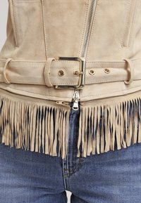 Giacca in suede beige con orlo frangiato, cintura con fibbia dorata e zip frontale, indossata su jeans in denim blu.