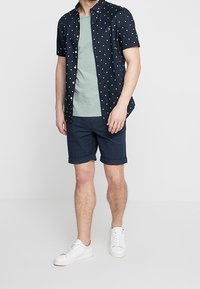 Marinblå kortärmad skjorta med vita prickar, grön t-shirt under, marinblå shorts och vita sneakers. Avslappnad passform, knäppt design.