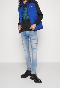 Chaleco inflado azul sobre un sudadera negra con un gráfico turquesa, combinado con jeans claros rasgados y zapatos negros.