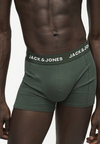 Ανδρικός κορμός με πράσινα boxer briefs Jack & Jones, με εμφανείς κοιλιακούς μύες και σκούρο τόνο δέρματος, τα χέρια χαλαρά στο πλάι.