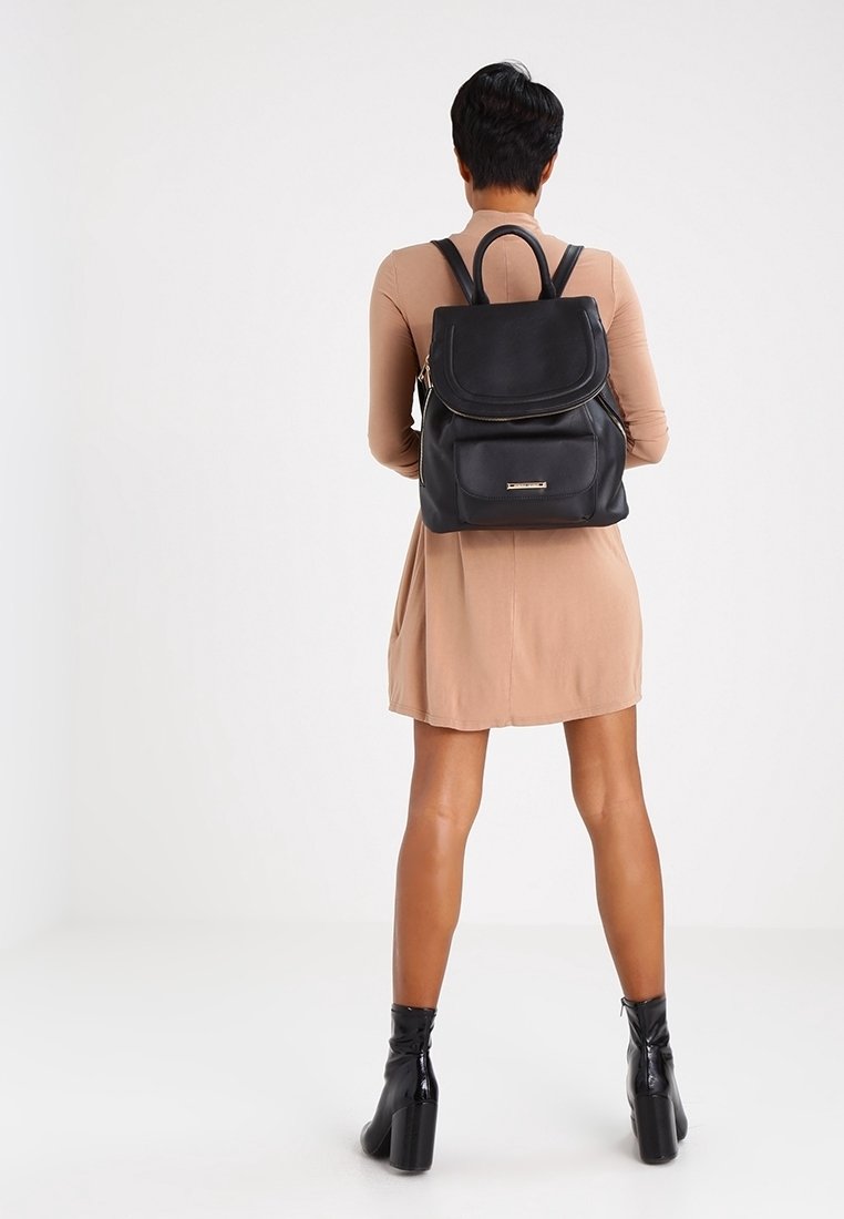 Femme en robe beige et bottines noires portant un sac à dos en cuir noir, debout devant un fond blanc uni.
