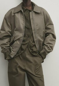 Olijfgroene bomberjack met een gladde textuur, rits aan de voorkant en grote kraag, gecombineerd met een bijpassend overhemd met knopen en een broek.
