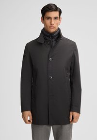 Cappotto nero con colletto alto e chiusura a bottoni, dotato di tasche laterali e un tessuto texturizzato. Lo strato interno è visibile al colletto.
