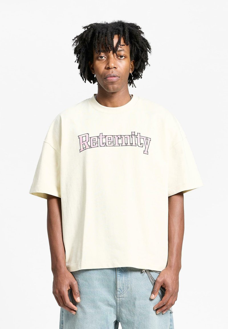 Kremfarget oversized t-skjorte med "Reternity" tekst i rosa. Korte ermer, rund hals, myk stoff, avslappet passform. Dongerishorts synlig.