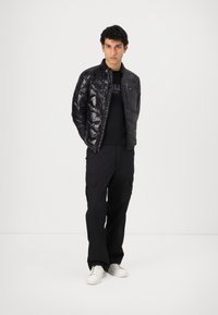 Giubbotto a sacco nero con finitura lucida, chiusura con zip e colletto alto, indossato sopra una t-shirt nera e abbinato a pantaloni cargo neri e sneakers bianche.