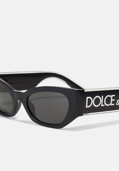 Occhiali da sole neri con una montatura spessa e lenti scure. Presentano un audace logo bianco sulla asta, evidenziato da un dettaglio in accento argentato.