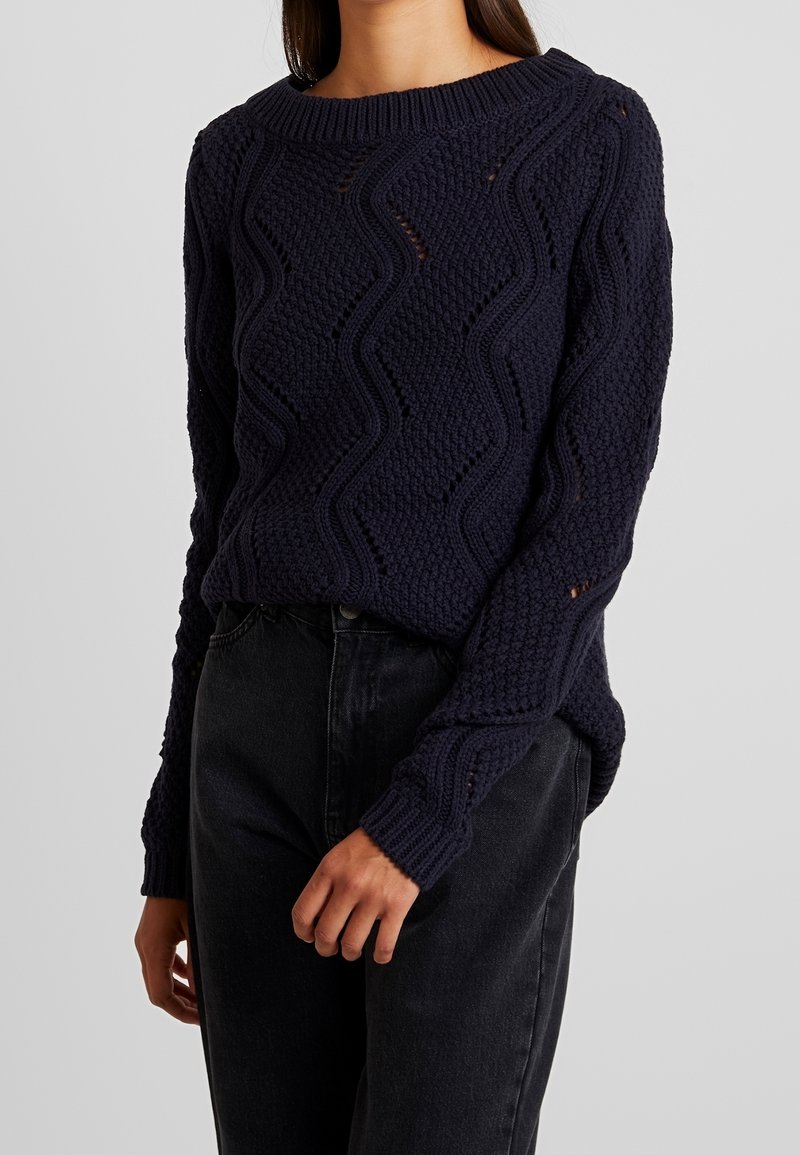 Femme portant un pull en maille bleu marine avec des motifs en zigzag et losanges, associé à un jean noir taille haute, sur un fond uni.