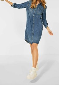 Kvinna som bär en blå denimklänning med knappar och vita högskaftade sneakers, balanserande på ett ben med armarna utsträckta mot en enkel bakgrund.