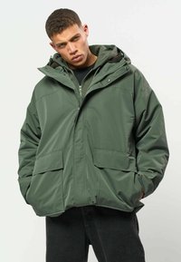 Jeune homme portant une grande veste à capuche vert olive avec les mains dans les poches, debout devant un fond clair uni.