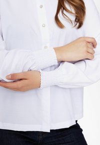 Camicia bianca con bottoni, caratterizzata da maniche a sbuffo con polsini arricciati. Tessuto liscio con una vestibilità comoda, che mostra una sottile trama.