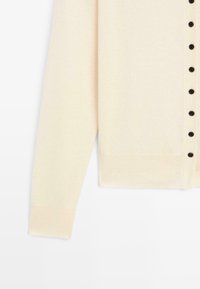Cardigan beige clair avec un devant à boutons noir, une texture douce et des manches longues. Coupe simple avec des poignets côtelés.
