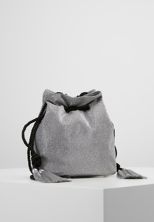 Sac à cordon texturé gris avec bretelles en corde noire torsadée et pompons, présenté sur une plateforme blanche sur fond uni.