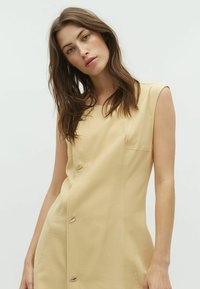 Abito beige senza maniche con un design aderente, caratterizzato da una tasca frontale e dettagli metallici in forma di bottoni lungo il lato. Tessuto morbido.