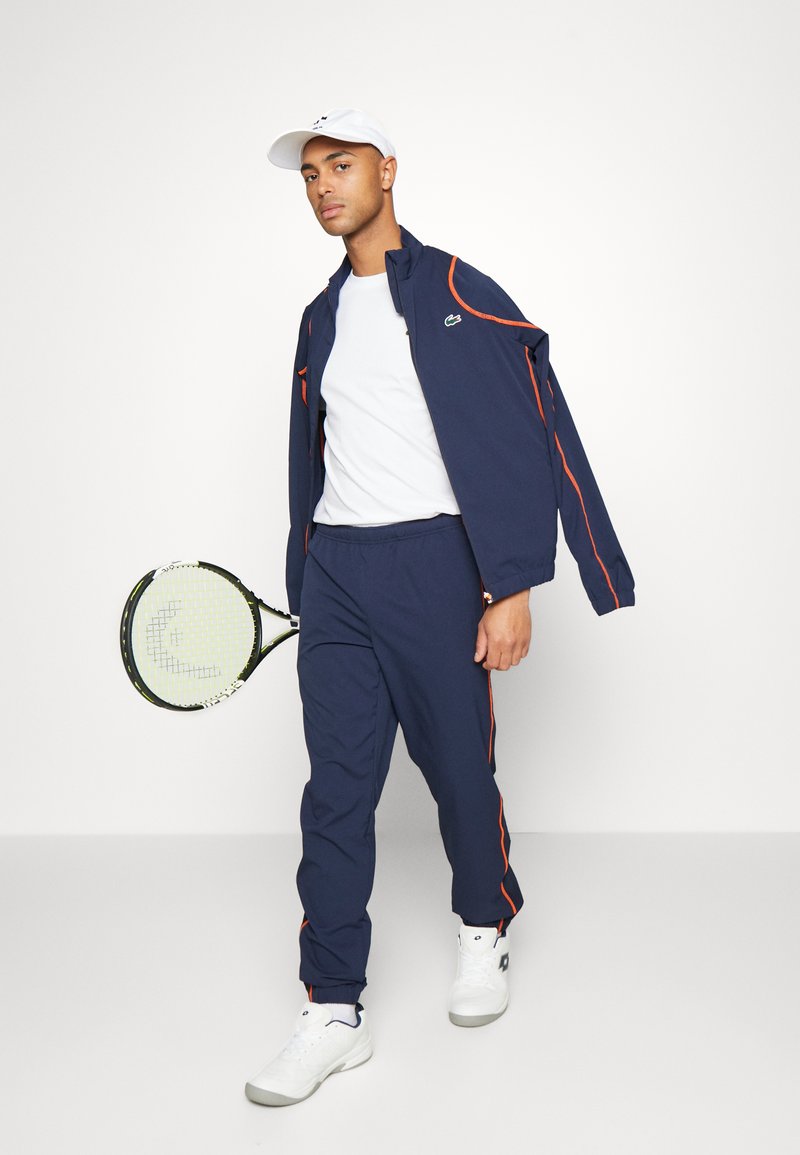Man in marine blauwe trainingsoutfit met oranje accenten, witte sneakers en witte pet, houdend een tennisracket, staand tegen een effen witte achtergrond.