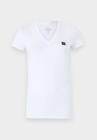 Camiseta de algodón blanca con cuello en V; de mangas cortas; presenta un pequeño parche de logo negro en el pecho; textura suave, diseño ajustado.