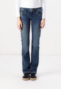 Jean en denim bleu avec une coupe droite, comportant cinq poches et une fermeture par bouton. Porté avec des baskets noires et un haut blanc à manches longues.