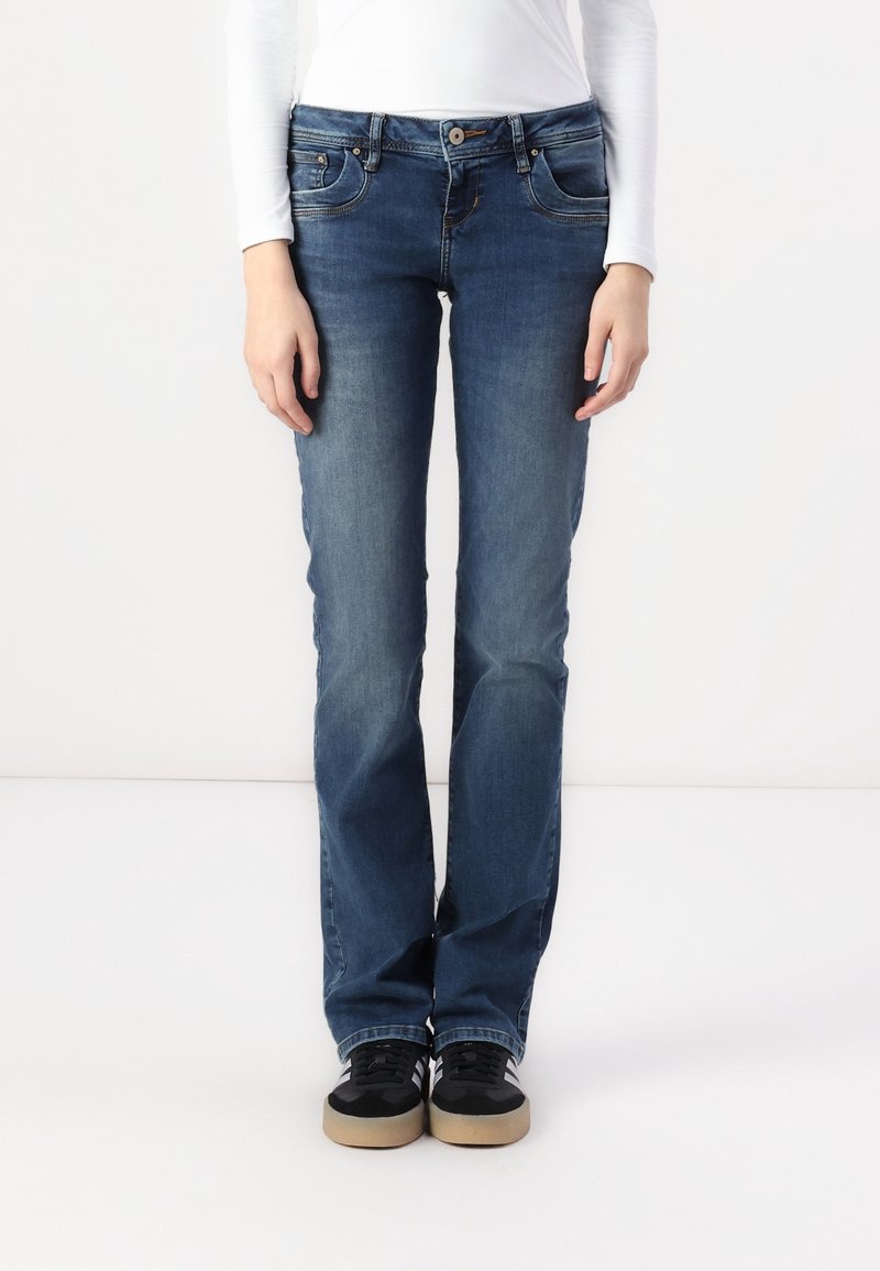 Jean en denim bleu avec une coupe droite, comportant cinq poches et une fermeture par bouton. Porté avec des baskets noires et un haut blanc à manches longues.