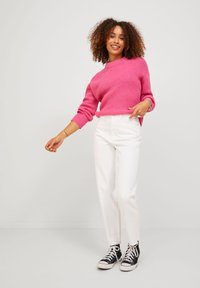 Pink knitted sweater with long sleeves, paired with white high-waisted pants and black canvas sneakers. Background is neutral.