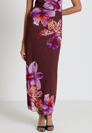Femme portant une jupe maxi bordeaux avec de grands imprimés floraux violets et orange et des talons ouverts noirs, debout devant un fond uni.