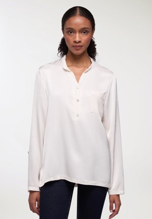 Blusa bianca a maniche lunghe con apertura a metà bottone, tasca sul petto e tessuto liscio e leggero. L'orlo è leggermente curvo.