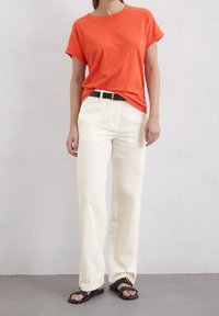 Orange kortærmet t-shirt parret med løstsiddende, cremefarvede bukser og sorte sandaler. Outfitet har et sort bælte.