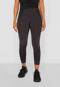 Leggings pretos feitos de tecido elástico, com painéis contornados e um detalhe do logótipo na perna esquerda, combinados com ténis de desporto pretos.