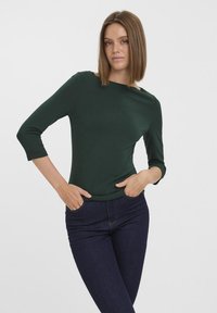 Top ajustat de culoare verde închis, cu mâneci trei sferturi și decolteu rotund, realizat dintr-un material neted. Purtați cu blugi negri, mâinile în buzunare.
