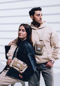 Borsa a tracolla beige con dettagli testurizzati e una chiusura. Giacca di jeans indossata sopra un felpa. Jeans grigi e un orologio visibili su un modello maschile.