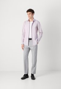 Helles rosa langärmliges Shirt, das über einem weißen T-Shirt getragen wird, kombiniert mit hellgrauen Hosen und schwarzen Schuhen. Einfaches Design, schlanker Schnitt.