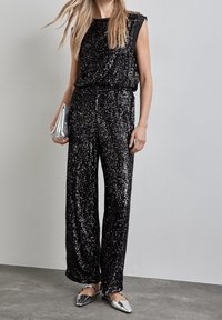 Czarny kombinezon z cekinami o luźnym kroju, z bezrękawnikowym fasonem i ściągniętą talią. W komplecie z metalicznymi srebrnymi balerinami i kopertówką.