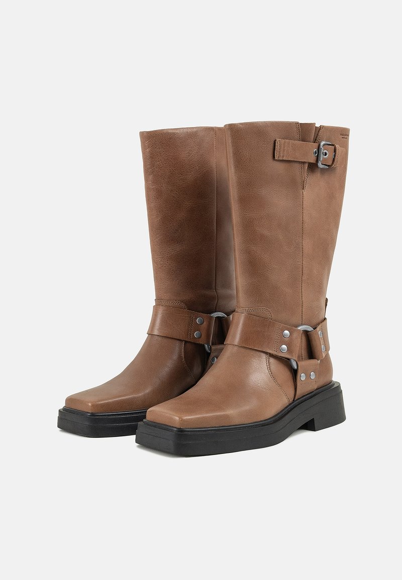 Botas de cuero marrón hasta la rodilla con punta cuadrada, detalles de hardware plateado y una suela de goma negra gruesa, con un detalle de hebilla en la correa.