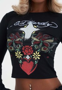 Schwarzes, tailliertes Longsleeve-Oberteil mit buntem Kreuz, Rosen, Blumen und Herz-Grafik sowie "Ed Hardy"-Text, getragen von einer Person mit Bauchnabelpiercing.