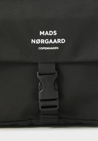 Svart stoffryggsekk med toppklaff, med plastspenne og hvit merkevaretekst: "MADS NØRGAARD COPENHAGEN."