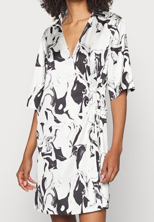Femme portant une robe portefeuille à motif abstrait en noir et blanc, manches aux coudes et col, debout les bras détendus.