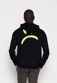 Felpa nera realizzata in tessuto morbido, con un grande motivo di sorriso giallo sul retro, dotata di cappuccio con coulisse e polsini rigati.