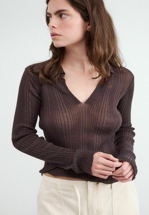 Jonge vrouw met schouderlang bruin haar, gekleed in een aansluitende donkerbruine geribde V-hals trui en lichte broek, kijkt opzij.