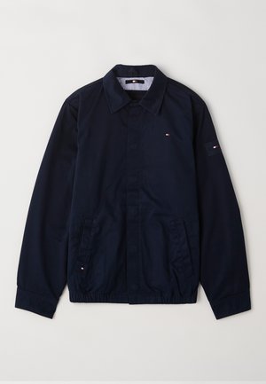 Tumš zilā jaka, izgatavota no kokvilnas, ar pogu aizdari, punktveida apkakli, sānu kabatām un logo uz krūškurvja un piedurknes. Strukturēta auduma.