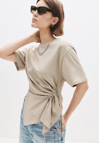 Kvinde iført en beige kortærmet bluse med sidelukninger sikret med en ring, lyseblå jeans, solbriller og en sølv kædedyse, der poserer med hånden på nakken.