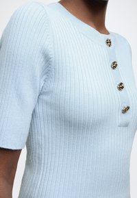 Ελαφρώς γαλάζιο ριμπ πλεκτό τοπ με κοντά μανίκια, με στρογγυλεμένη λαιμόκοψη και χρυσά διακοσμητικά κουμπιά κατά μήκος της γραμμής κουμπώματος.