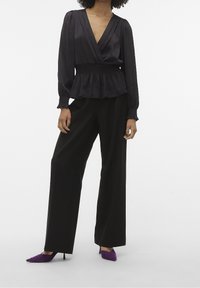 Svart, lättvikts omslagblus med plisserad textur, långa ärmar och smockad midja, tillsammans med högmidjade svarta byxor och lila klackar.