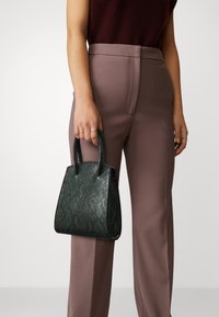 Vihreä käärmekuvioinen käsilaukku, jossa on jämäkkä muoto, tukevat kaksinkertaiset kahvat ja kiiltävä viimeistely, pidetty vaaleanruskeiden housujen päällä.