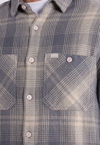 Chemise en flanelle à carreaux grise et beige. Comprend deux poches poitrine, des boutons clairs et un col. Tissu texturé.
