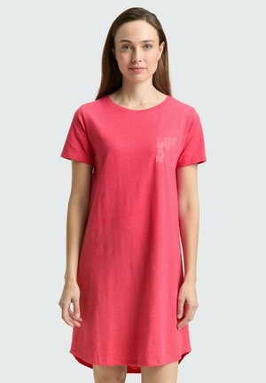 Vrouw met lang bruin haar die een koraalroze casual jurk met korte mouwen draagt, staand tegen een effen lichte achtergrond.