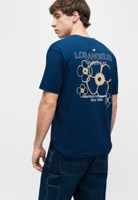 Νεαρός άνδρας που φοράει μπλε σκούρο μπλουζάκι με μπεζ λουλουδάτα σχέδια και το κείμενο "LOS ANGELES City park tour" στην πλάτη, συνδυασμένο με σκούρο μπλε τζιν παντελόνι.