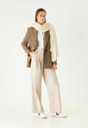 Brauner Blazer über einem weißen Hemd, kombiniert mit beigen, weit geschnittenen Hosen und einem cremefarbenen Schal. Weiße Sneaker mit schwarzen Akzenten runden den Look ab.