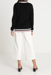 Svart stickad tröja med kontrasterande vita och röda ränder på muddar och nederkant, kombinerad med vida vita byxor och svarta klackskor.