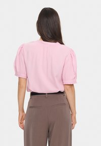 Rosa Kurzarmbluse mit puffigen Ärmeln und gerafftem Rückendetail, kombiniert mit braunen Hosen mit Gesäßtaschen und einem Gürtel.