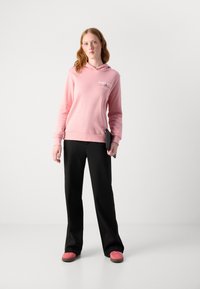Sudadera rosa claro con un logo en la parte delantera, combinada con pantalones anchos negros y zapatos rosas. La tela parece suave con un ajuste relajado.