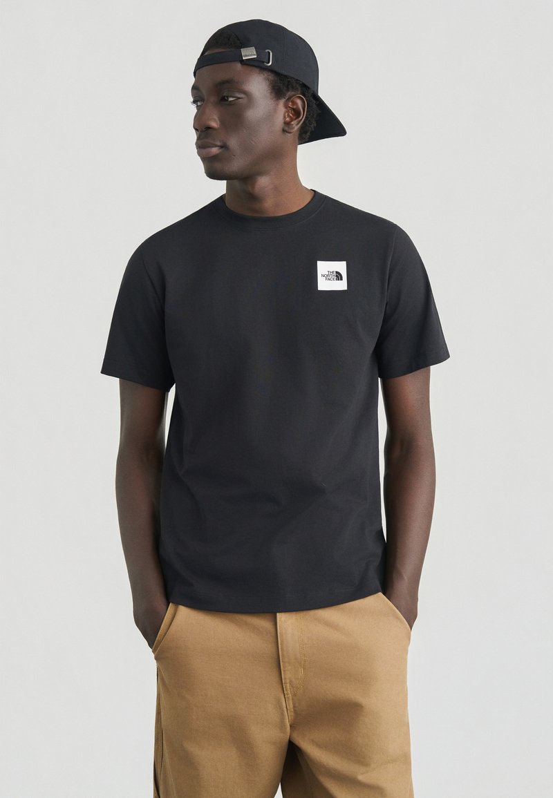 Μαύρο βαμβακερό μπλουζάκι με κοντά μανίκια, με λευκό τετράγωνο λογότυπο στο αριστερό στήθος. Συνδυασμένο με ανοιχτό καφέ παντελόνι.