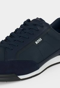 Baskets bleu marine à lacets avec tissu texturé et empiècements en suède, semelle blanche et noire, et logo "BOSS" sur le côté.